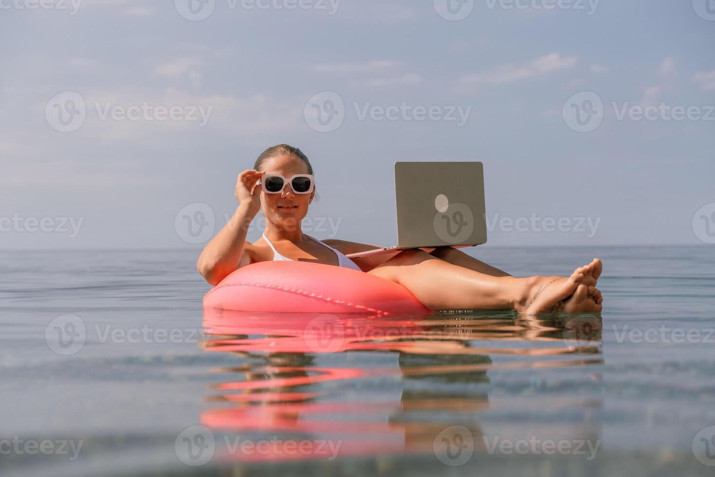 A woman is floating on a pink inflatable raft in the ocean while holding a laptop. Concept of relaxation and leisure, as the woman is enjoying her time in the water while working on her laptop. photo