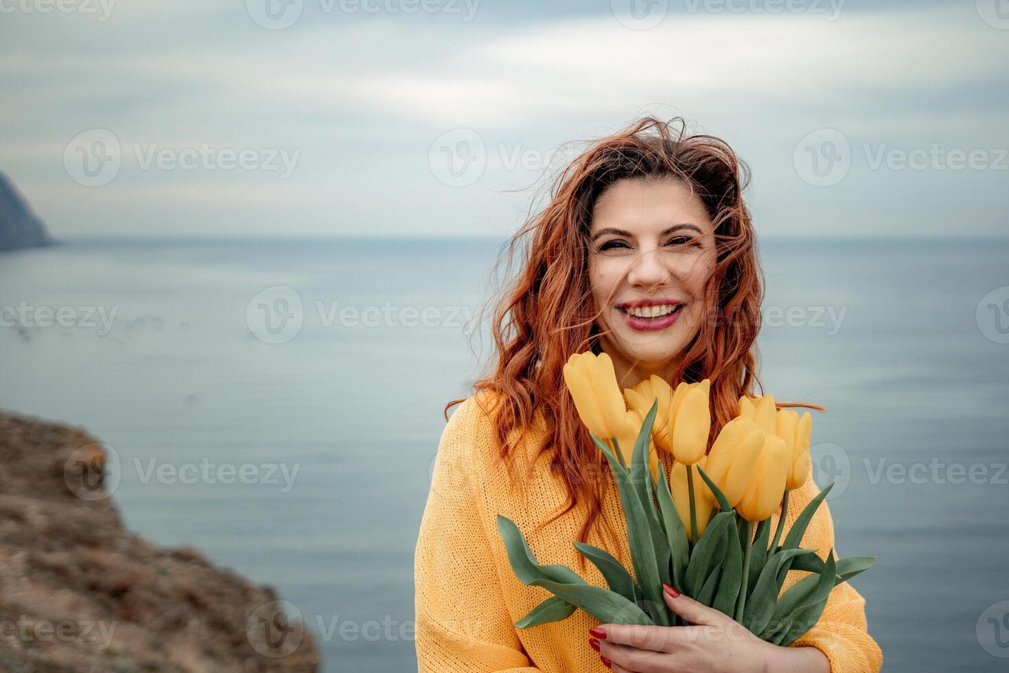 Portrait of a happy woman with hair flying in the wind against the backdrop of mountains and sea. Holding a bouquet of yellow tulips in her hands, wearing a yellow sweater photo