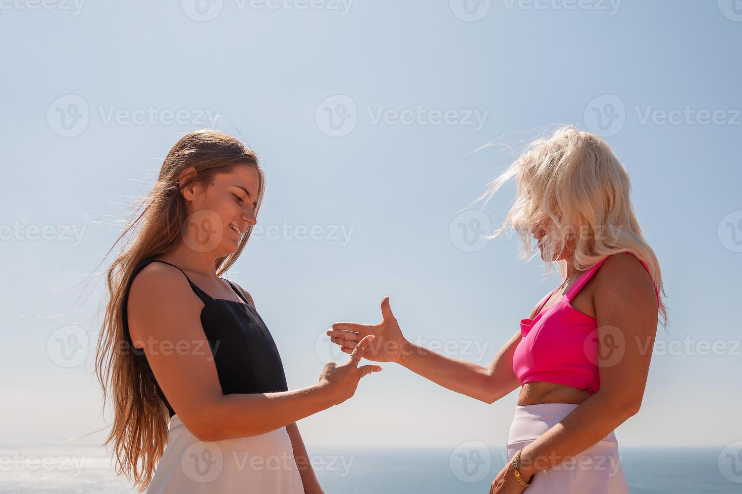 Two women are standing on a beach, one wearing a black tank top and the other wearing a pink top. They are both smiling and shaking hands. photo