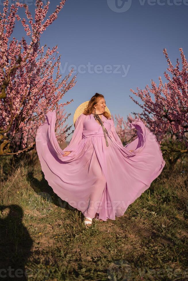 Woman blooming peach orchard. Against the backdrop of a picturesque peach orchard, a woman in a long pink dress and hat enjoys a peaceful walk in the park, surrounded by the beauty of nature. photo