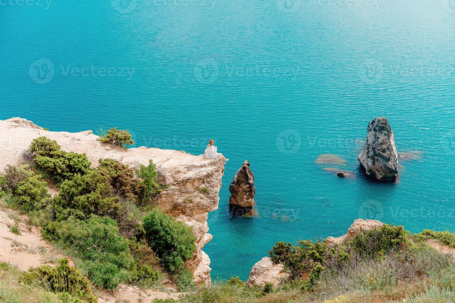 A woman sits on a rock overlooking the ocean. The water is blue and calm. The scene is peaceful and serene. photo