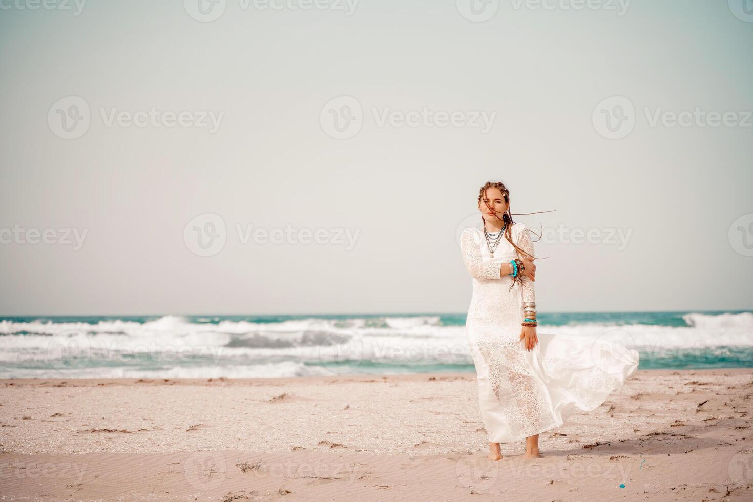 Model in boho style in a white long dress and silver jewelry on the beach. Her hair is braided, and there are many bracelets on her arms. photo