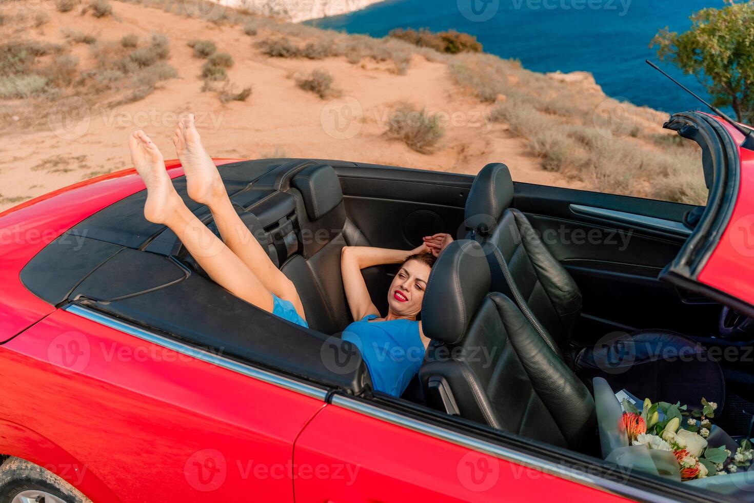 A woman is laying in the back of a red convertible car, with her feet up in the air. The car is parked on a cliff overlooking the ocean. Concept of freedom and relaxation. photo