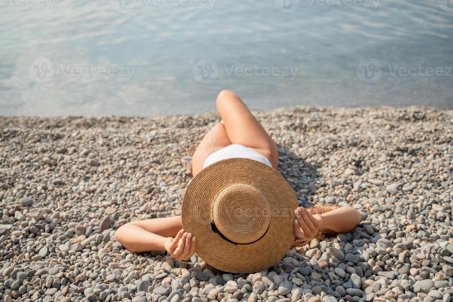 A woman is laying on the beach wearing a straw hat and a white bikini. She is enjoying the sun and the beach. photo