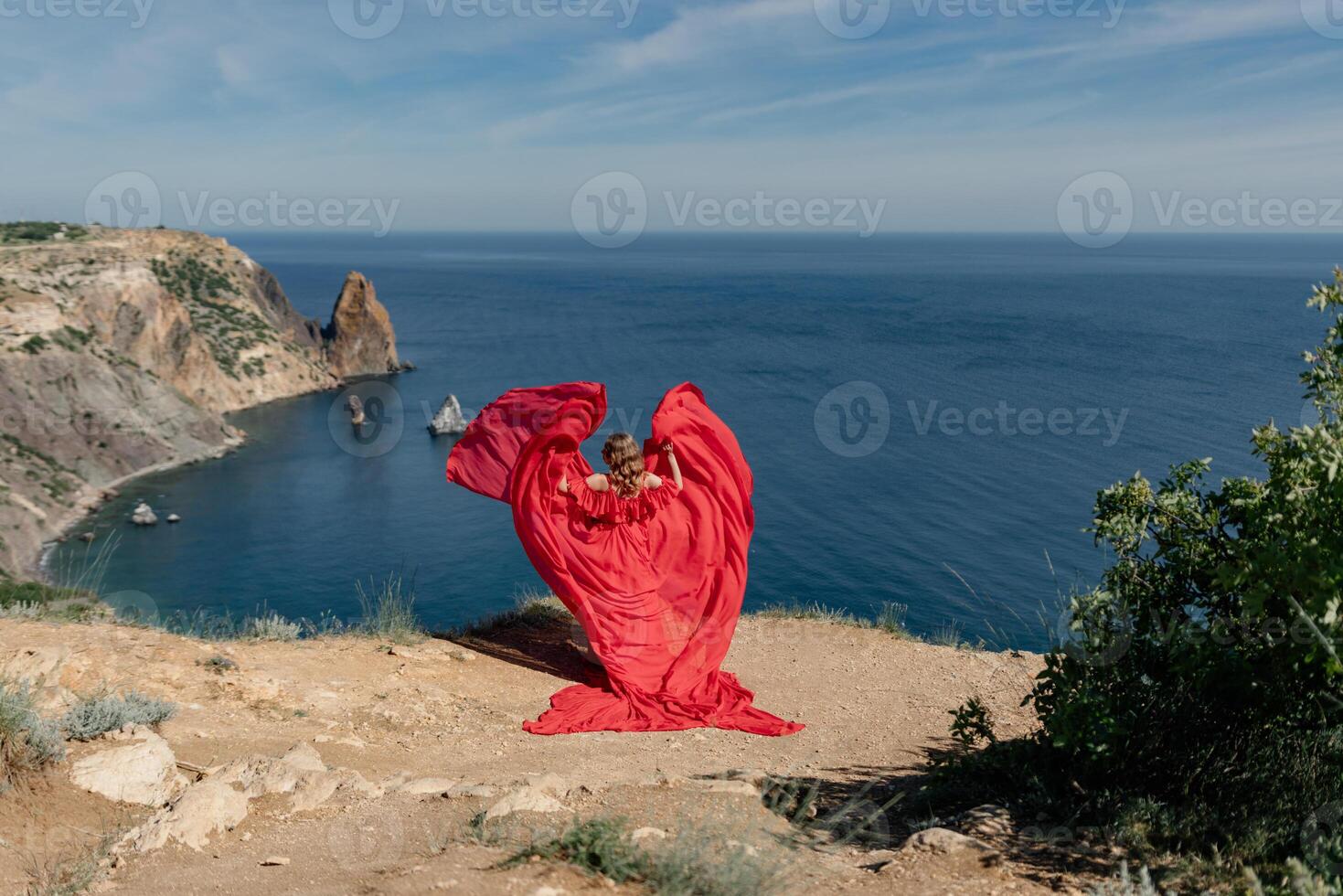 A woman in a red dress is standing on a rocky beach, with the ocean in the background. She is wearing a red dress and her hair is flowing in the wind. The scene is serene and peaceful. photo