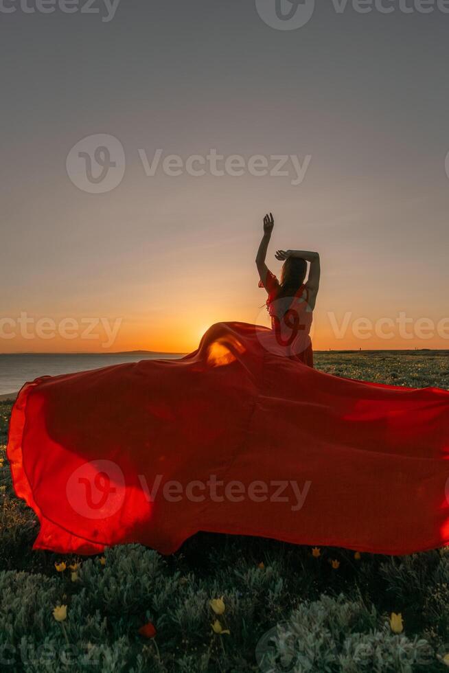 A woman in a red dress is standing in a field with the sun setting behind her. She is reaching up with her arms outstretched, as if she is trying to catch the sun. The scene is serene and peaceful. photo