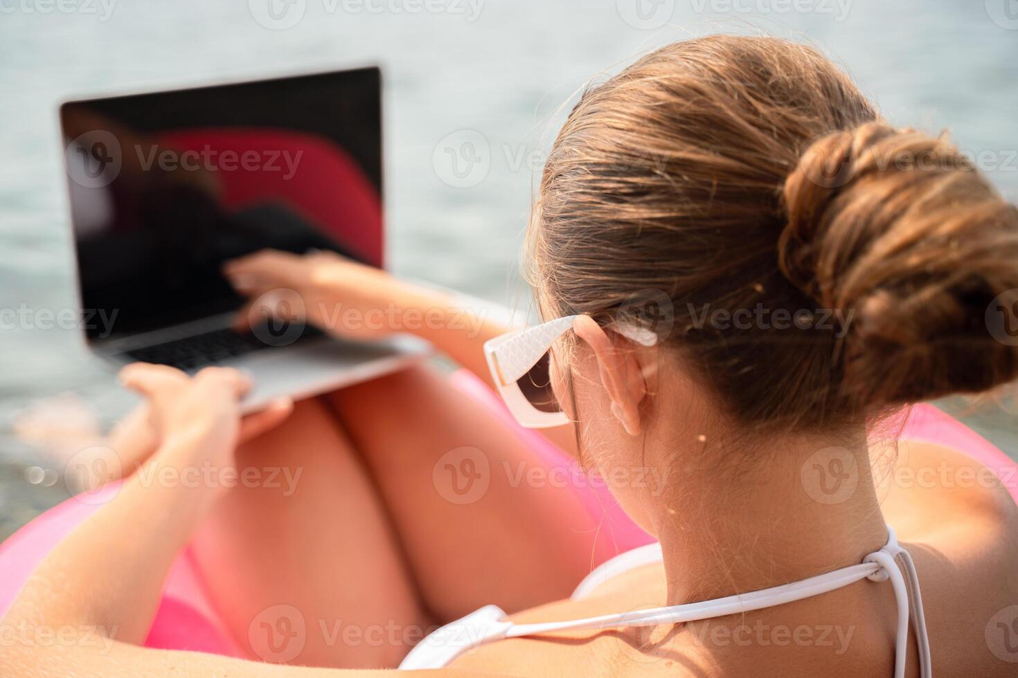 A woman is sitting in a pink inflatable raft with a laptop on her lap. She is wearing sunglasses and she is enjoying her time by the water. photo