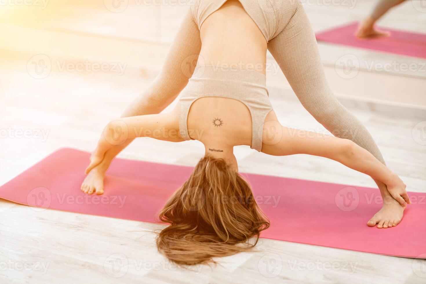 Young woman doing yoga in the gym. A girl with long hair and in a beige tracksuit stands in the Prasarita Padottanasana pose on a pink carpet. A woman performs Prasarita Padottanasana. photo