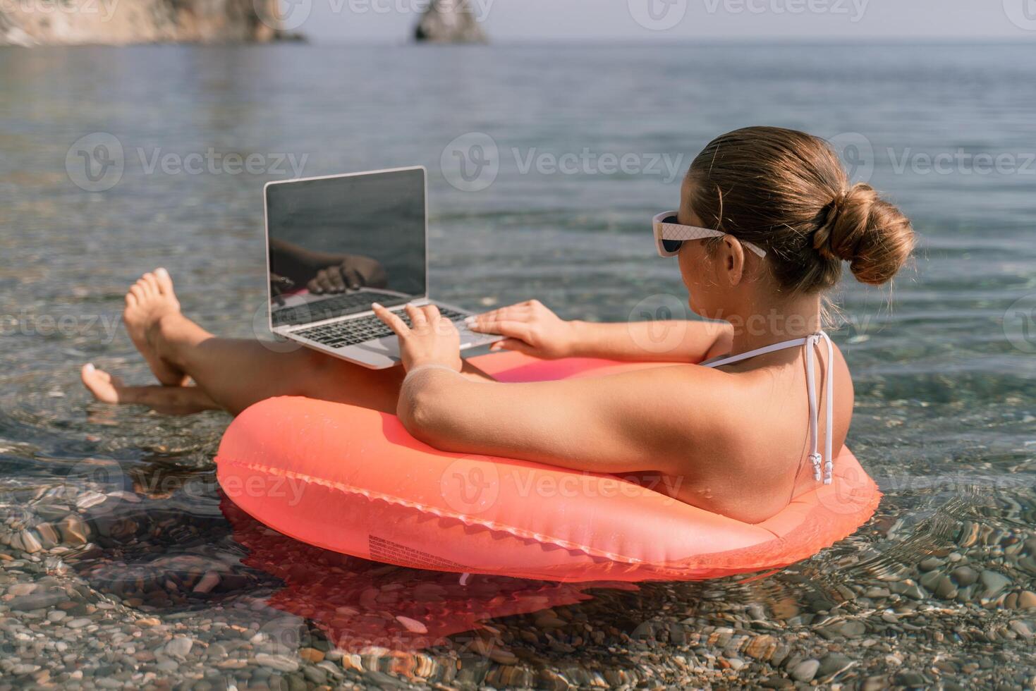 A woman is sitting on a pink inflatable raft in the water, using a laptop. Concept of relaxation and leisure, as the woman is enjoying her time by the water while working on her laptop. photo