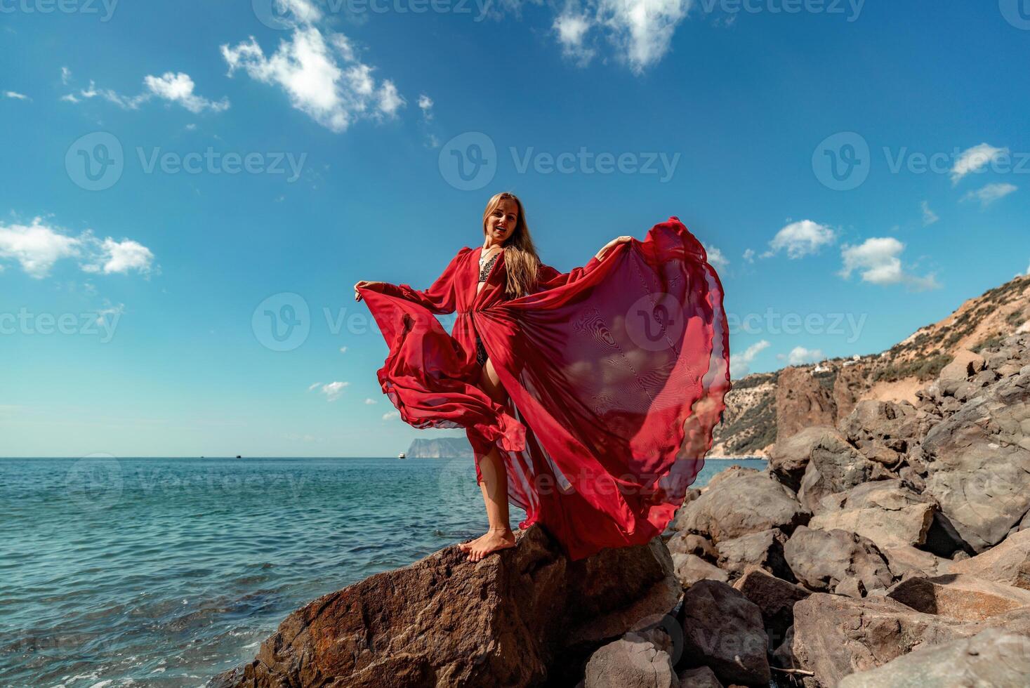 Red dress sea woman. A blonde with flowing hair in a long flowing red dress stands on a rock near the sea. Travel concept, photo session at sea