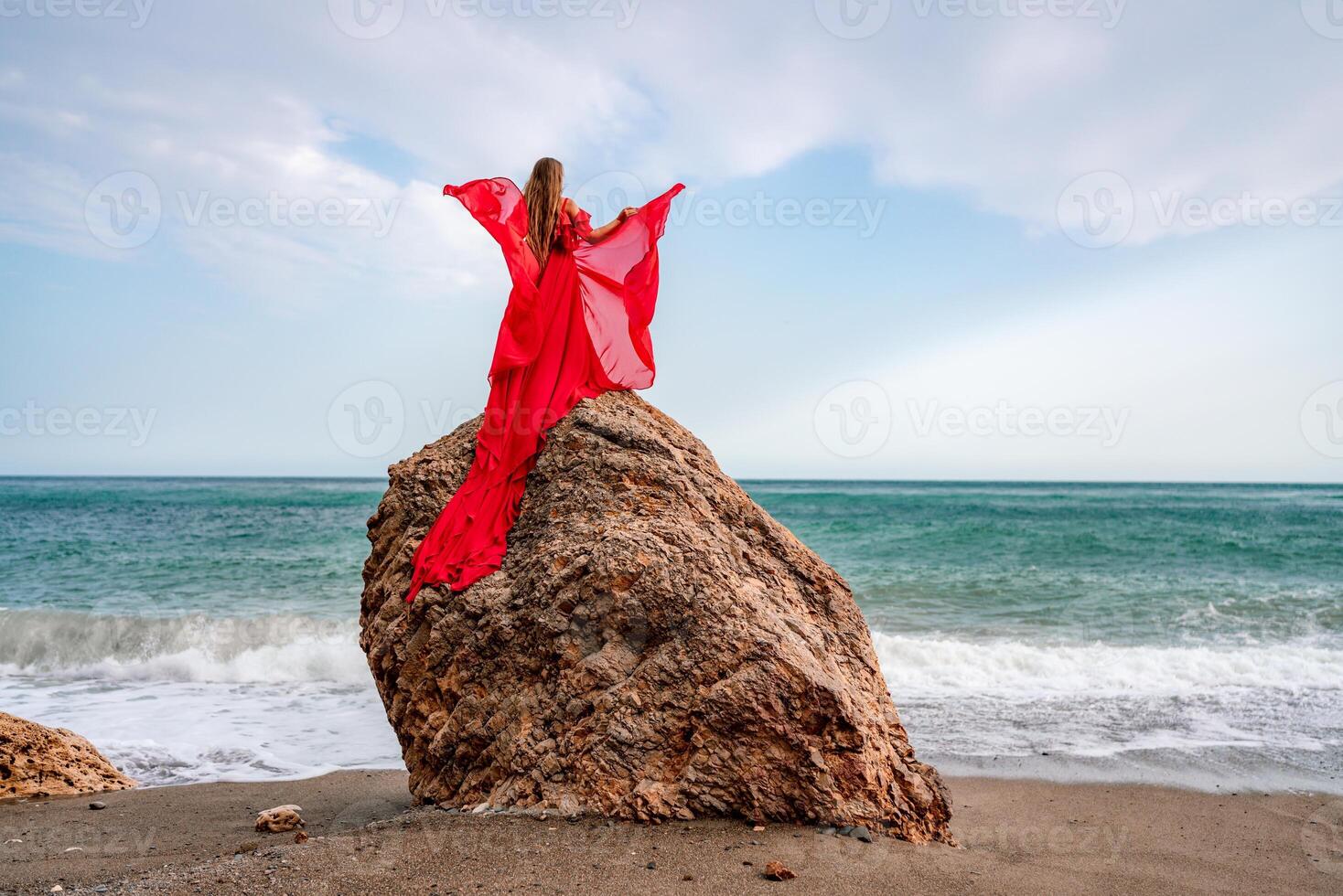 woman sea red dress. Woman with long hair on a sunny seashore in a red flowing dress, back view, silk fabric waving in the wind. Against the backdrop of the blue sky and mountains on the seashore. photo