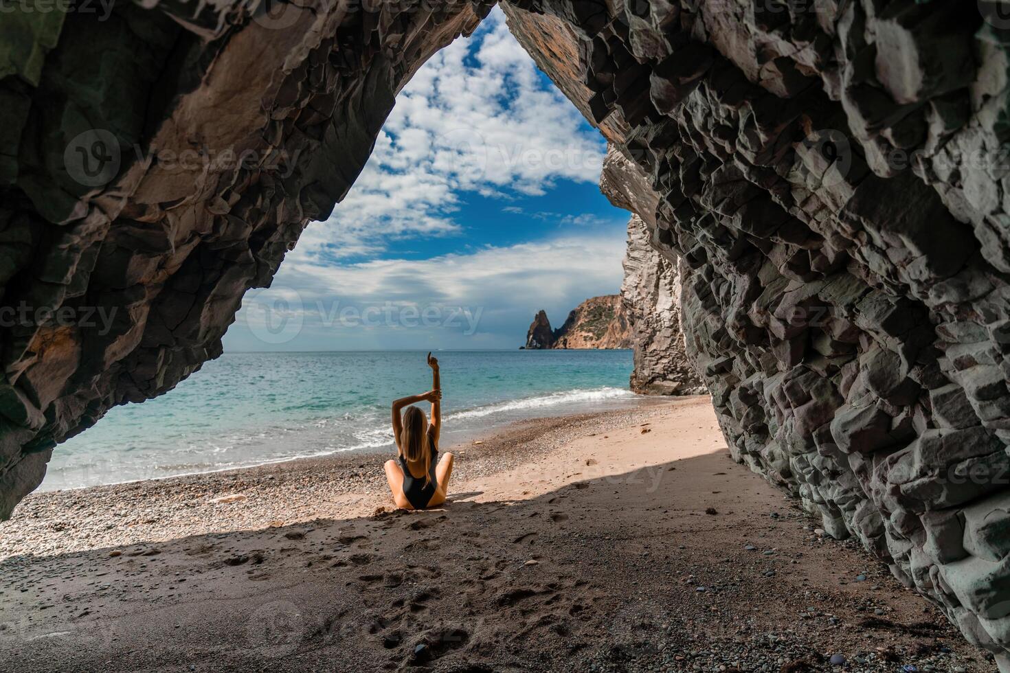 View of a woman in a black swimsuit from a sea cave Attractive woman enjoying the sea air sits on the beach and looks at the sea. photo