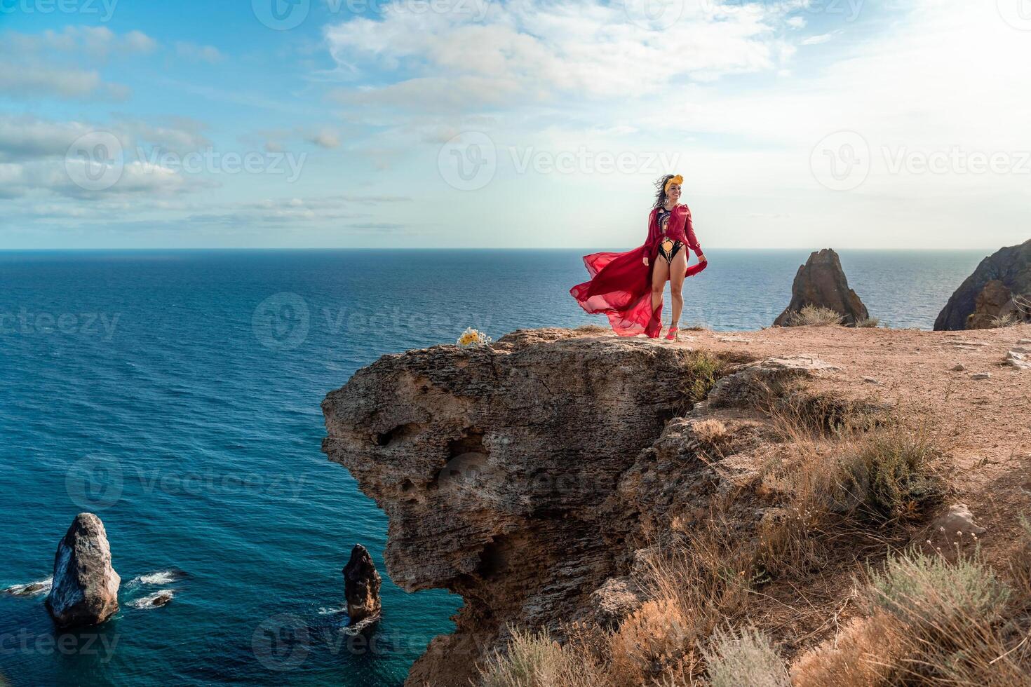 A woman in a red dress stands on a rocky cliff overlooking the ocean. The scene is serene and peaceful, with the woman's flowing dress adding a sense of grace and beauty to the landscape. photo
