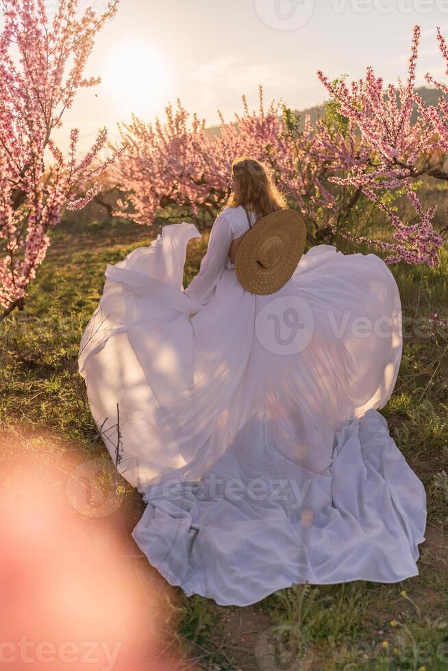 Woman blooming peach orchard. Against the backdrop of a picturesque peach orchard, a woman in a long white dress and hat enjoys a peaceful walk in the park, surrounded by the beauty of nature. photo