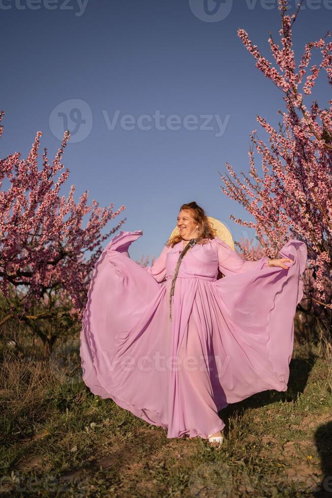 Woman blooming peach orchard. Against the backdrop of a picturesque peach orchard, a woman in a long pink dress and hat enjoys a peaceful walk in the park, surrounded by the beauty of nature. photo