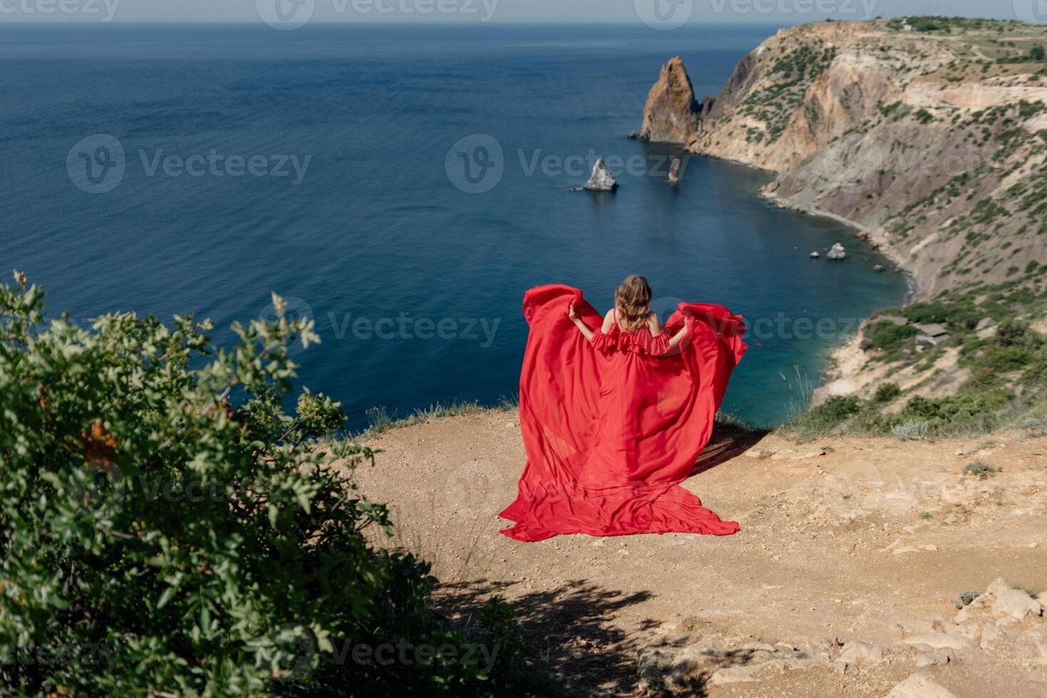 A woman in a red dress stands on a rocky beach, with the ocean in the background. The scene is serene and peaceful, with the woman's flowing dress adding a sense of grace and beauty to the landscape. photo