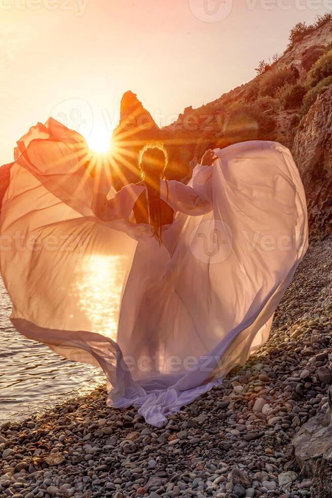 Mysterious woman silhouette long hair walks on the beach ocean water, sea nymph wind listens to the wave. Throws up a long white dress, a divine sunset. Artistic photo from the back without a face