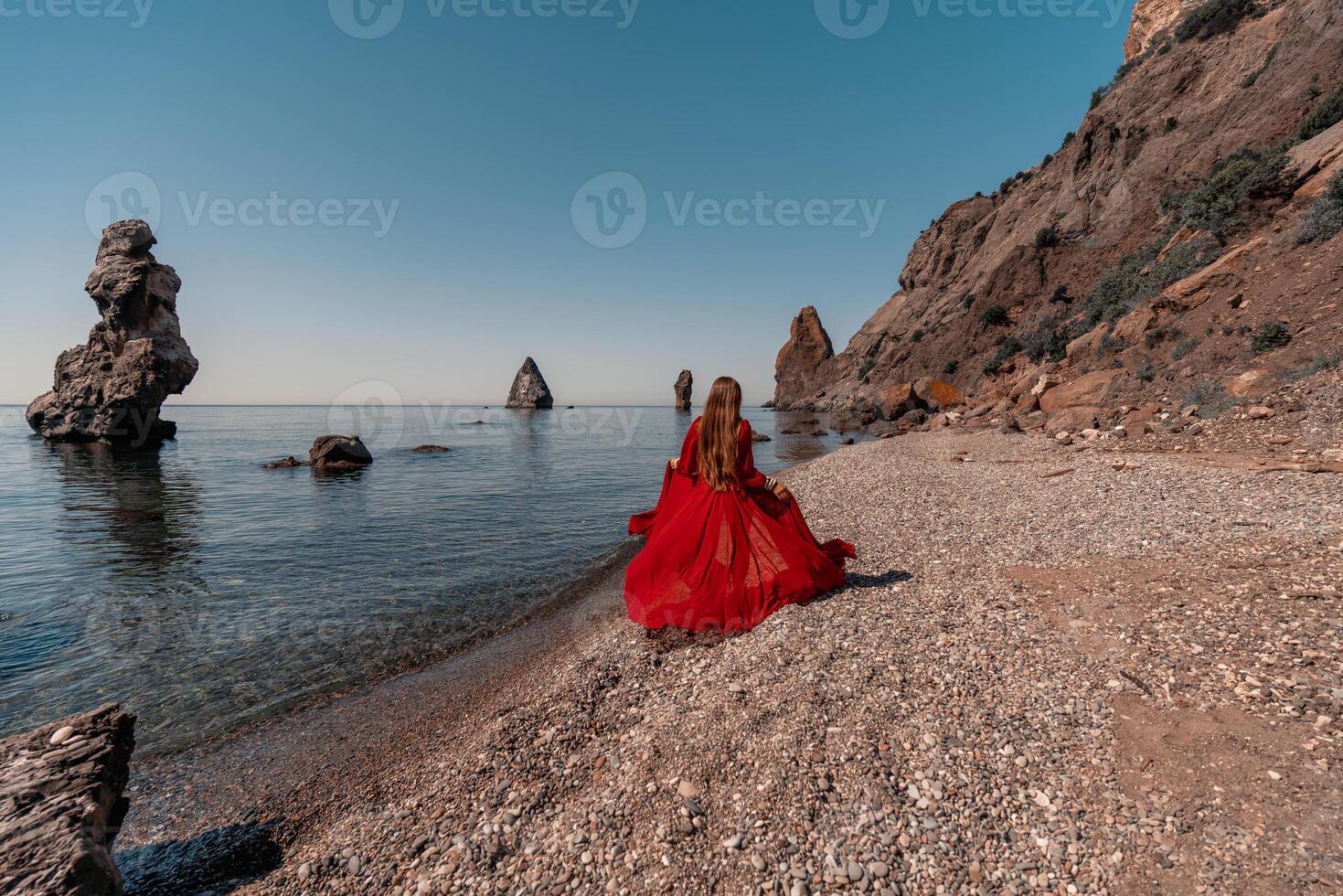 A woman in a red dress stands on a beach with a rocky shoreline in the background. The scene is serene and peaceful, with the woman's red dress contrasting against the natural elements of the beach. photo