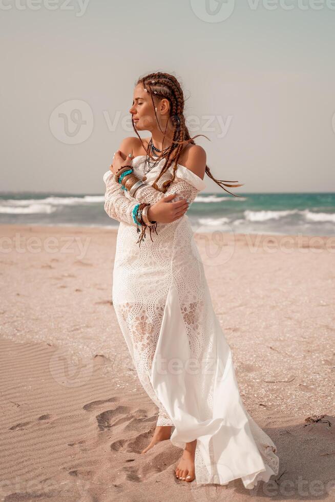 Model in boho style in a white long dress and silver jewelry on the beach. Her hair is braided, and there are many bracelets on her arms. photo