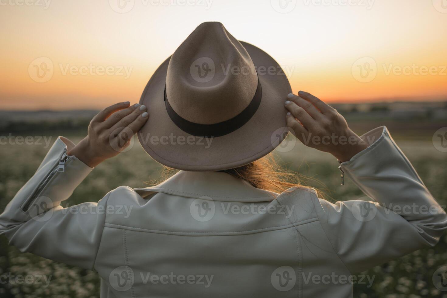 A woman wearing a hat and a white jacket is standing in a field. The woman is looking up at the sky, which is orange and pink. The scene has a peaceful and serene mood. photo