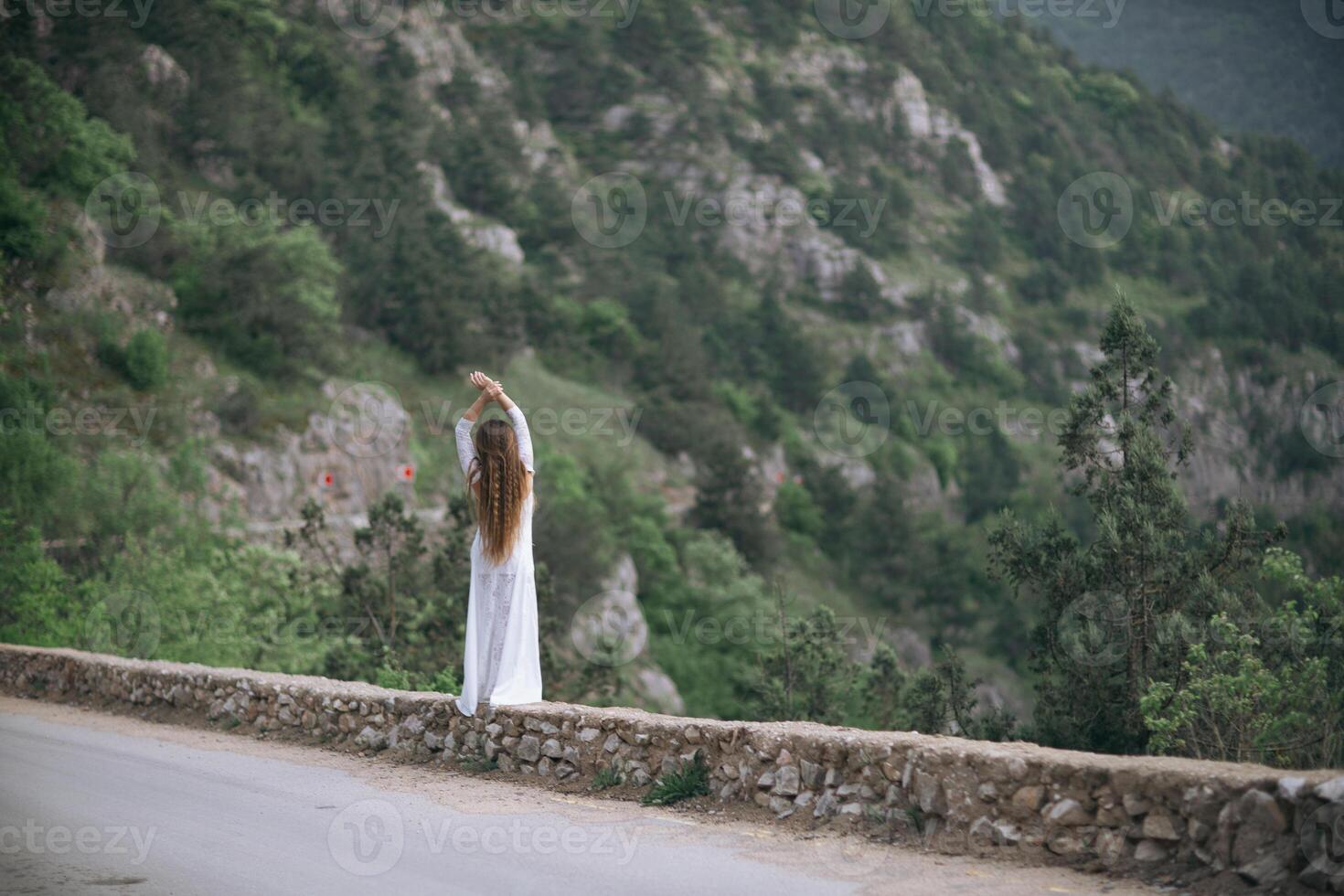 A woman stands on a stone wall overlooking a mountain. She is wearing a white dress and she is in a contemplative mood. photo