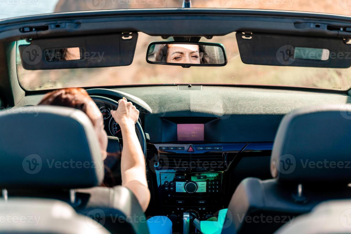 A woman is driving a convertible car with the top down. She is looking at her reflection in the rearview mirror. photo