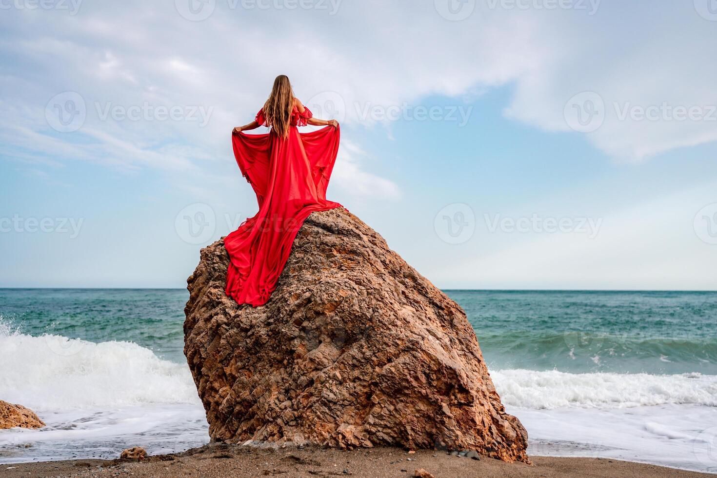 woman sea red dress. Woman with long hair on a sunny seashore in a red flowing dress, back view, silk fabric waving in the wind. Against the backdrop of the blue sky and mountains on the seashore. photo