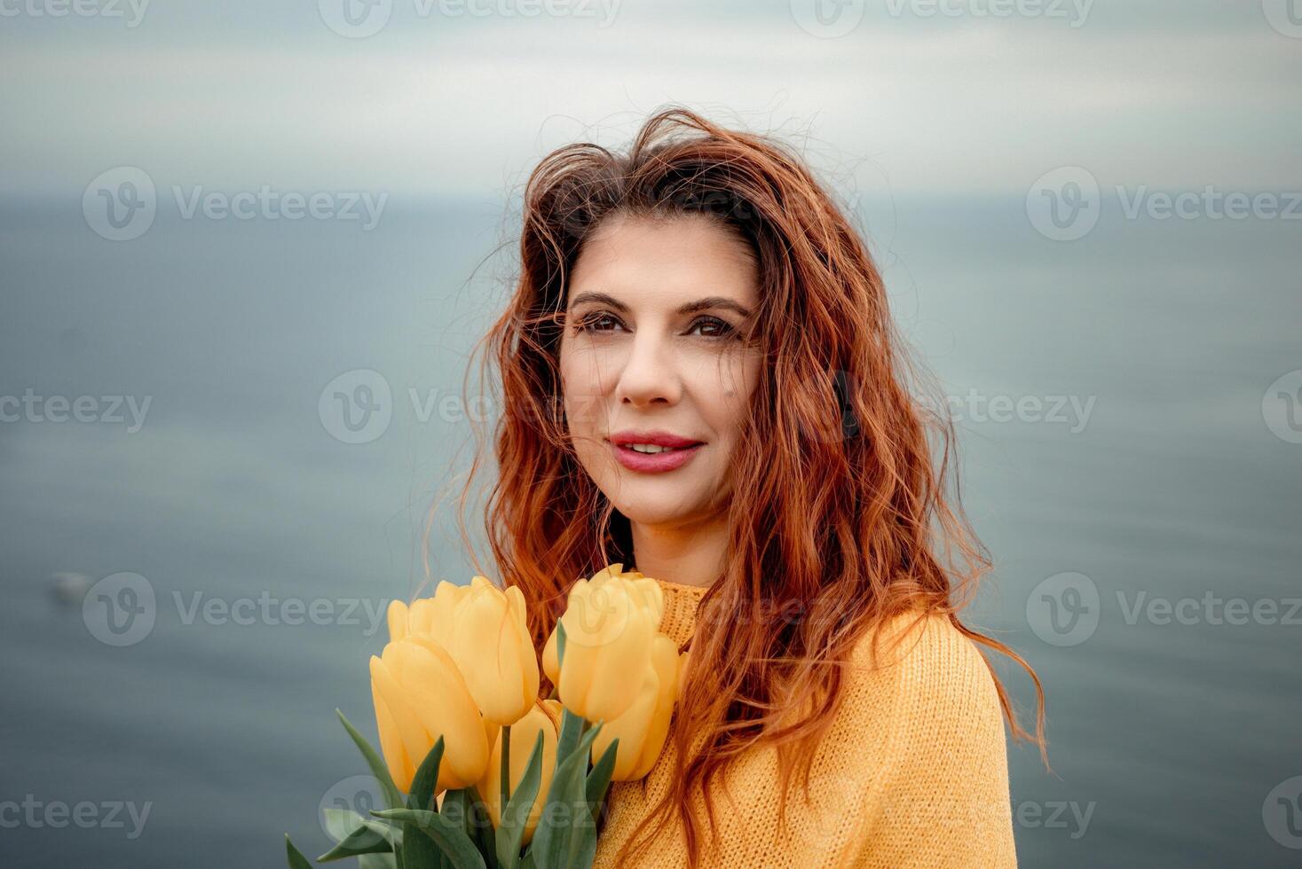 Portrait of a happy woman with hair flying in the wind against the backdrop of mountains and sea. Holding a bouquet of yellow tulips in her hands, wearing a yellow sweater photo