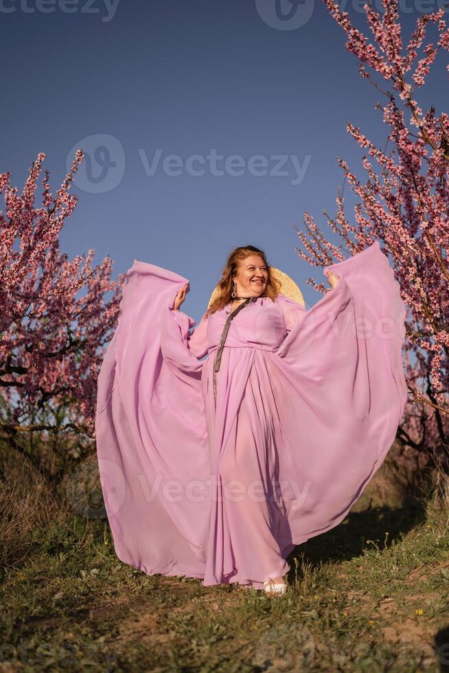 Woman blooming peach orchard. Against the backdrop of a picturesque peach orchard, a woman in a long pink dress and hat enjoys a peaceful walk in the park, surrounded by the beauty of nature. photo