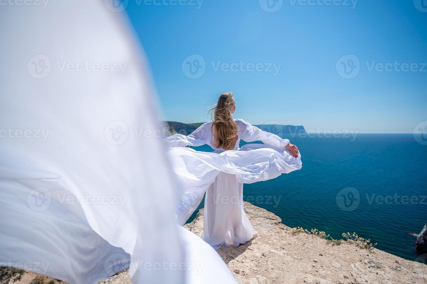 Blonde with long hair on a sunny seashore in a white flowing dress, rear view, silk fabric waving in the wind. Against the backdrop of the blue sky and mountains on the seashore. photo