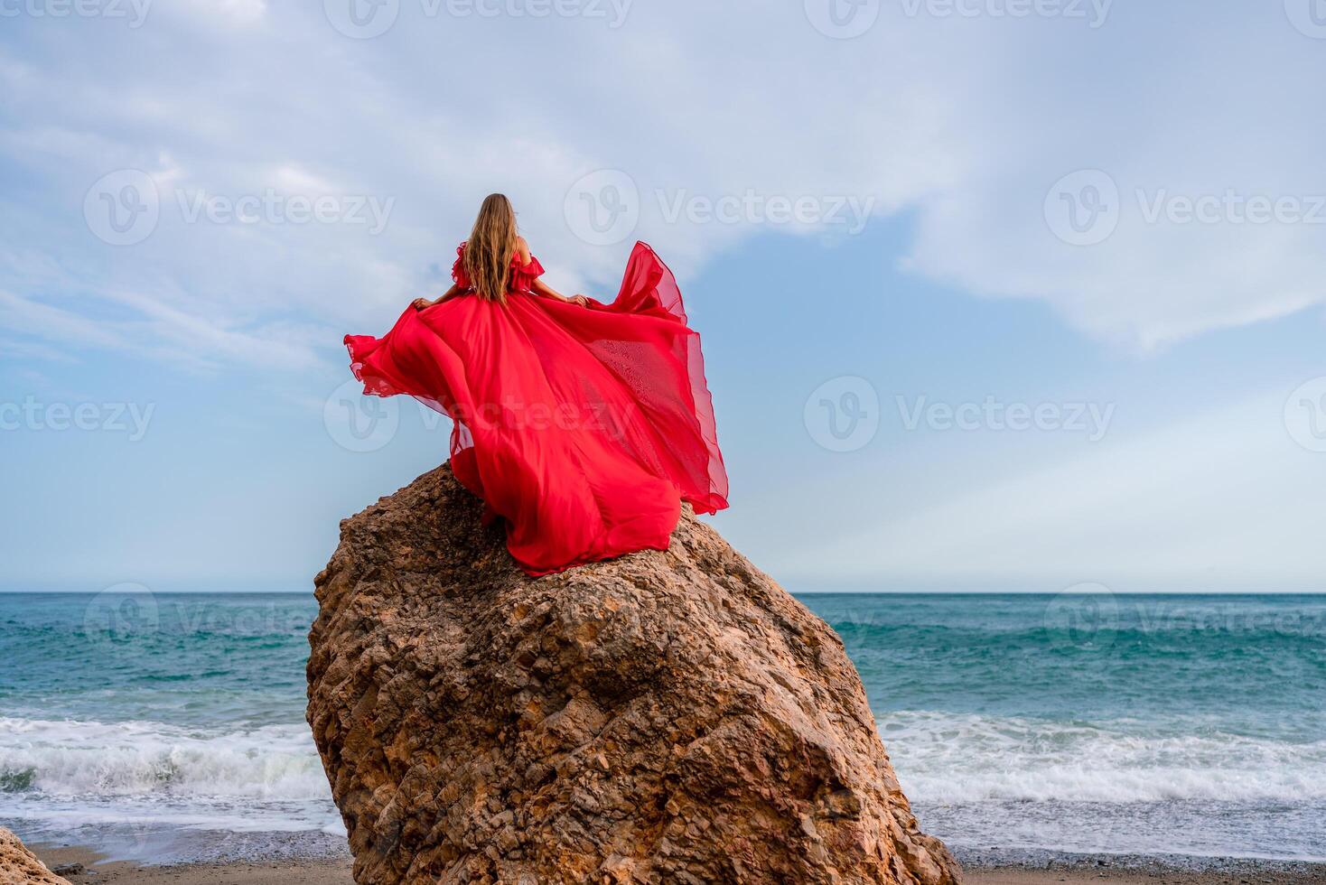 woman sea red dress. Woman with long hair on a sunny seashore in a red flowing dress, back view, silk fabric waving in the wind. Against the backdrop of the blue sky and mountains on the seashore. photo