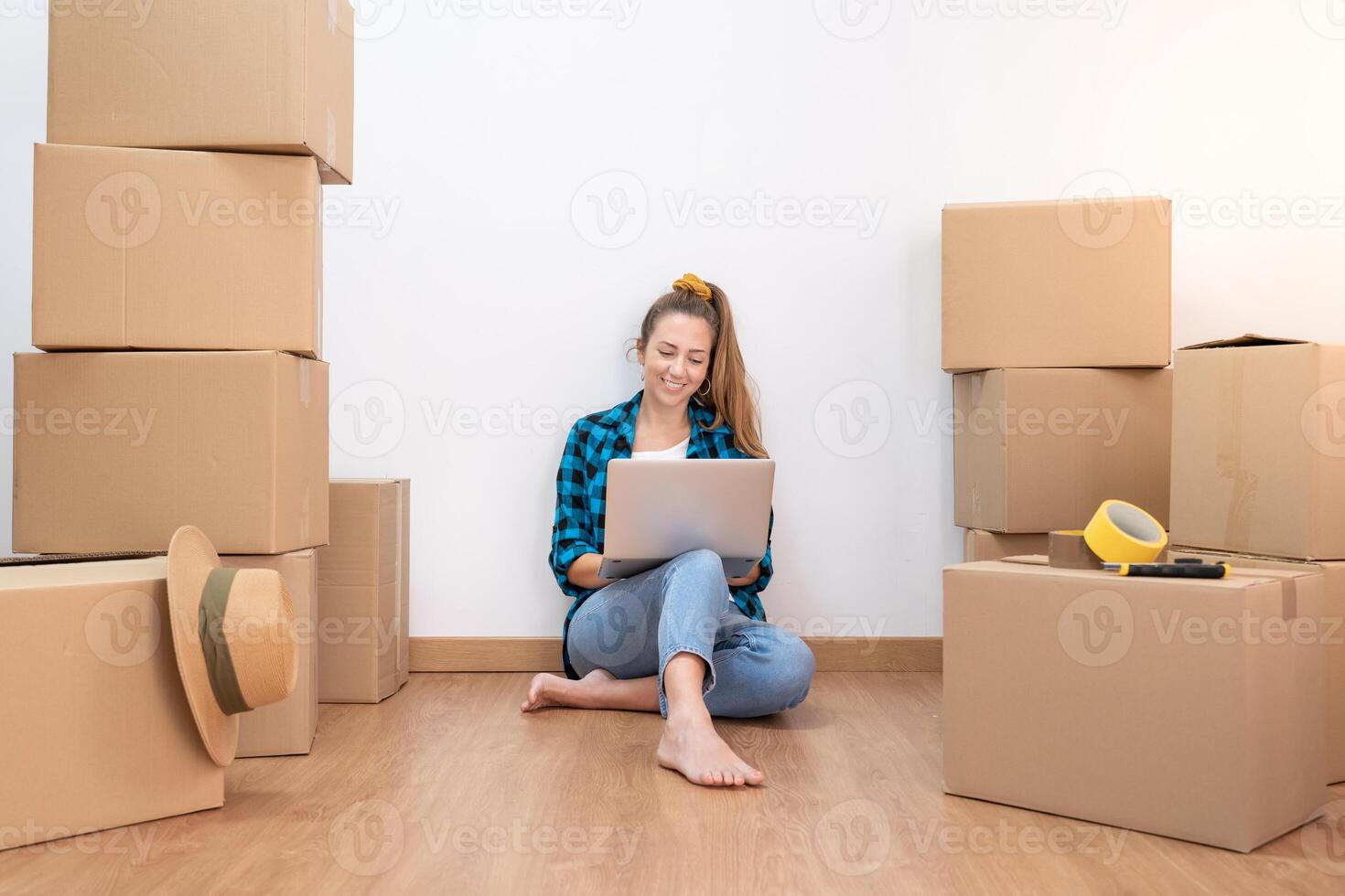 Young happy woman sitting on the floor with many boxes, working with computer in a new house. photo