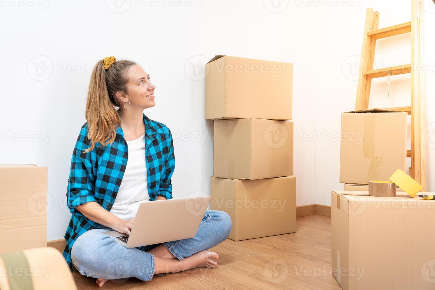 Young happy woman sitting on the floor with many boxes, working with computer in a new house. photo