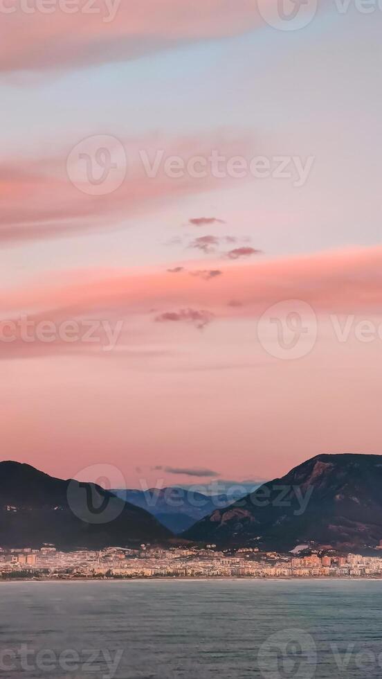 Pink clouds in evening sky over blue sea on background of city and mountains on horizon, vertical frame. photo