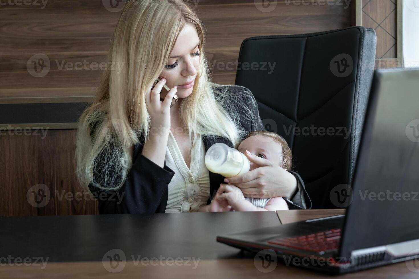 Young business woman feeds baby from bottle in office and talks on phone, concept of combining business and caring for newborn baby. photo