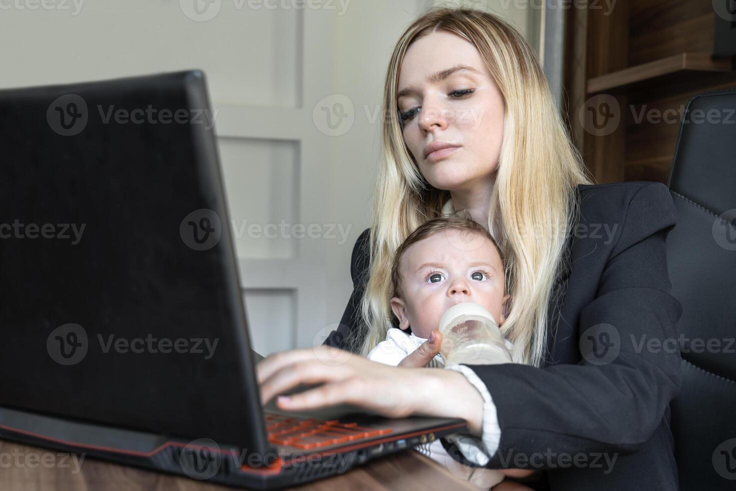 Concept of combining business and caring for a newborn child, young business woman feeds a baby from bottle in office, combining it with work on laptop. photo