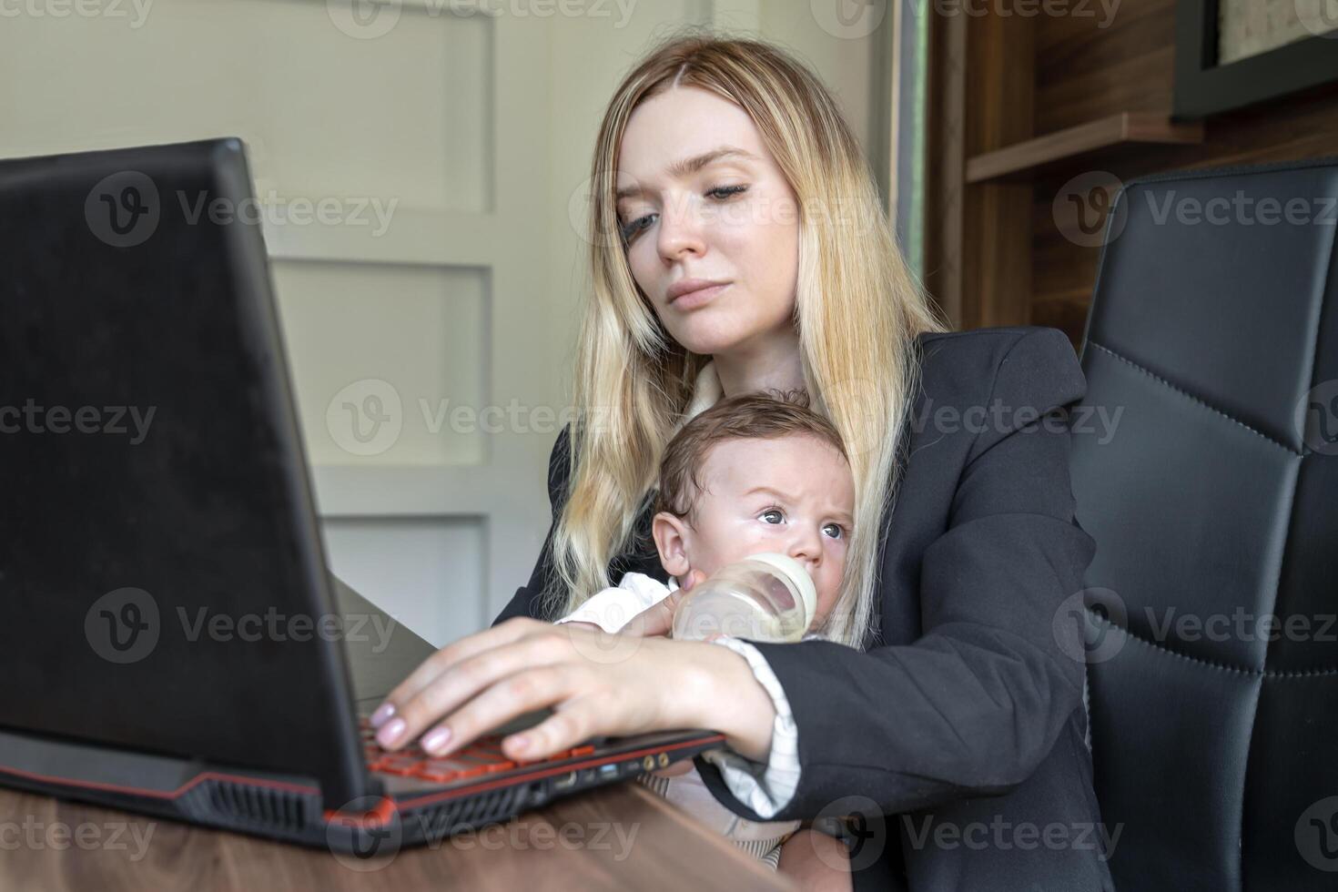 Concept of combining business and caring for newborn baby. young businesswoman holds baby in her arms, in office, combining this with working on laptop photo