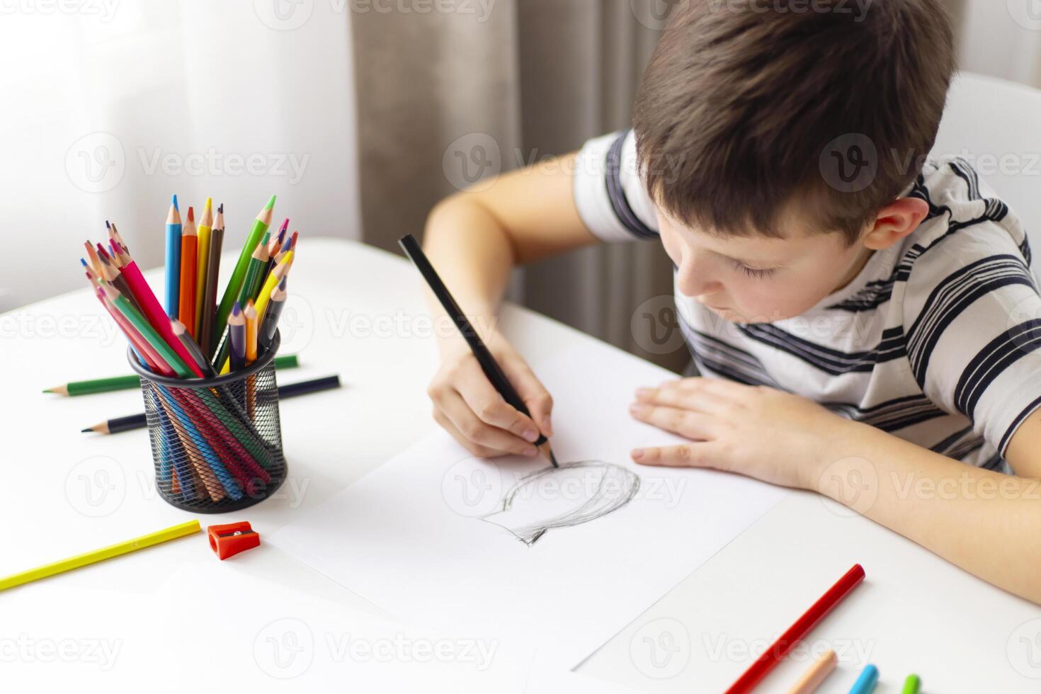 A child boy draws on white paper with colored pencils while sitting at a table photo
