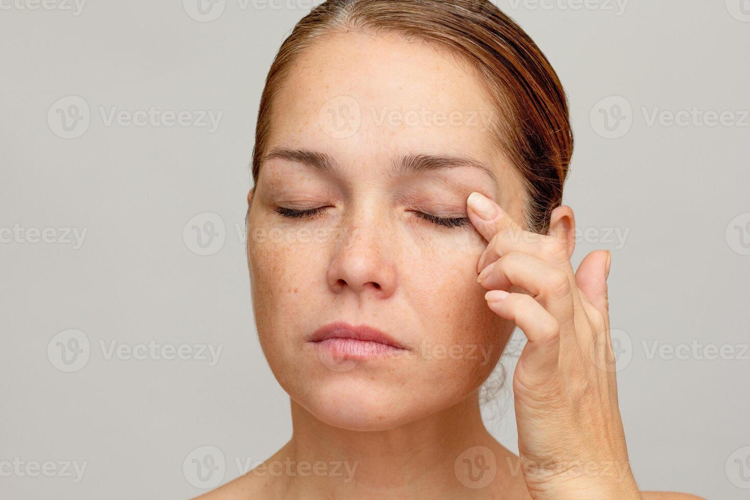 Middle aged caucasian woman of 40s with closed eyes holding finger on eyelid to show eyes aging on gray background photo
