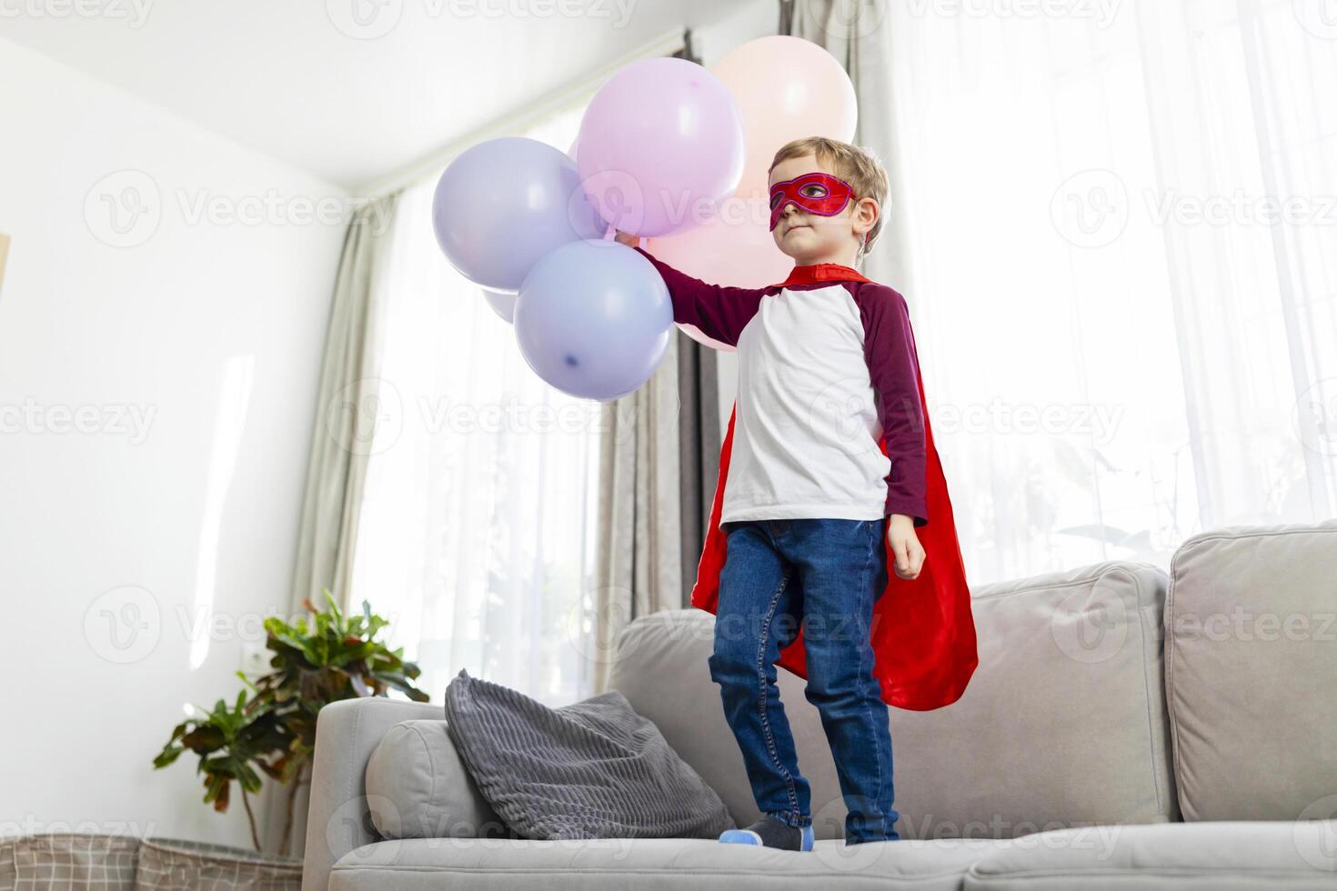 Boy in superhero costume with balloons standing on sofa photo