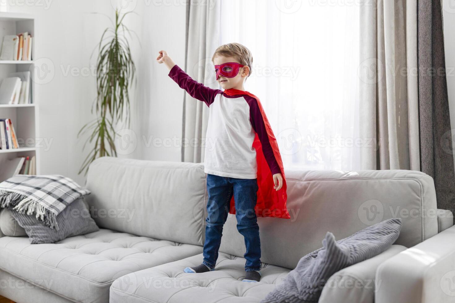 Child in superhero costume with red cape and mask standing on a sofa photo