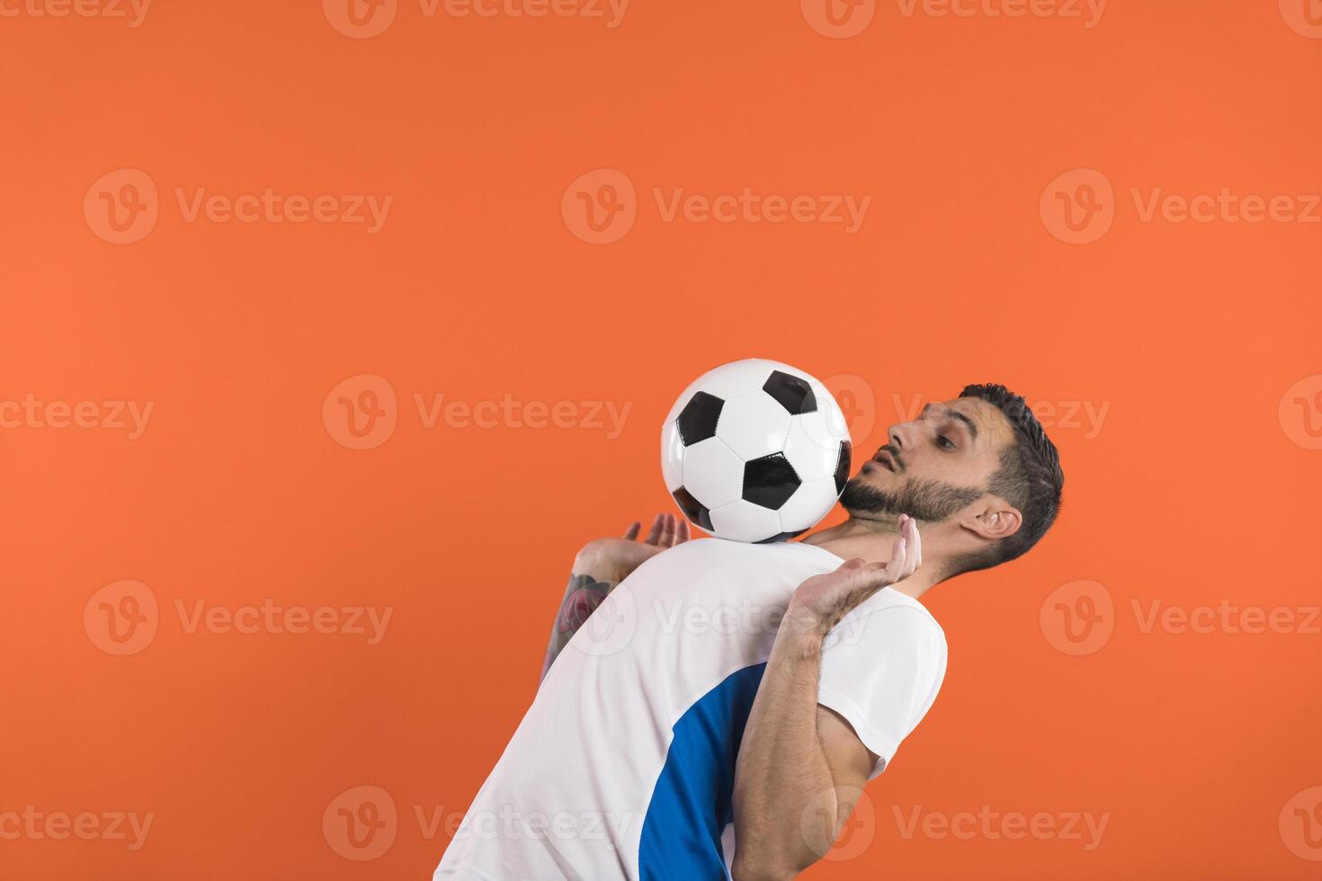 Concentrated football player in white blue shirt doing tricks with soccer ball, isolated on orange background photo