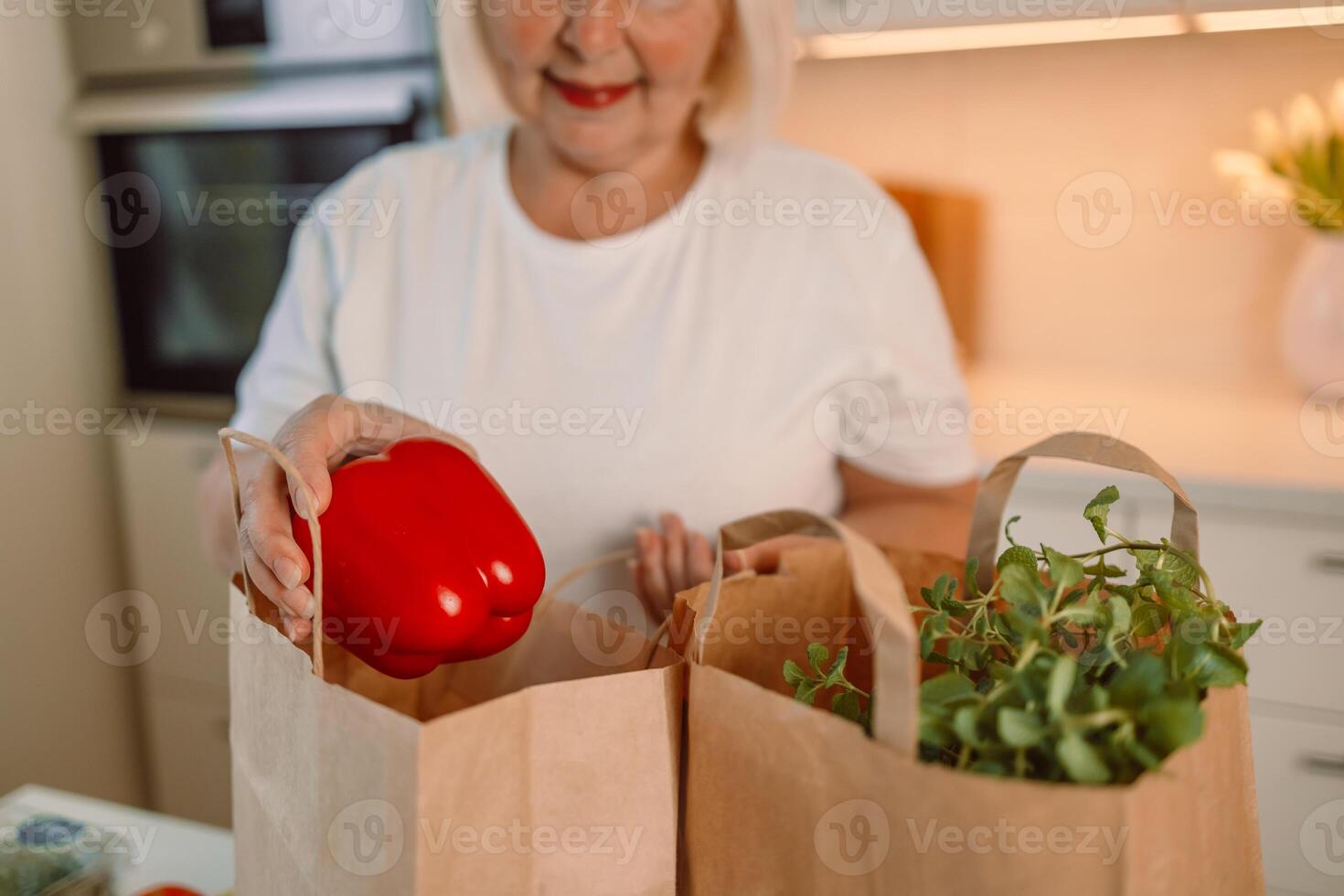 Happy Caucasian 60s woman sort vegetables from eco package in minimalist kitchen. Online buying food and grocery. Delivery service and mobile app. photo