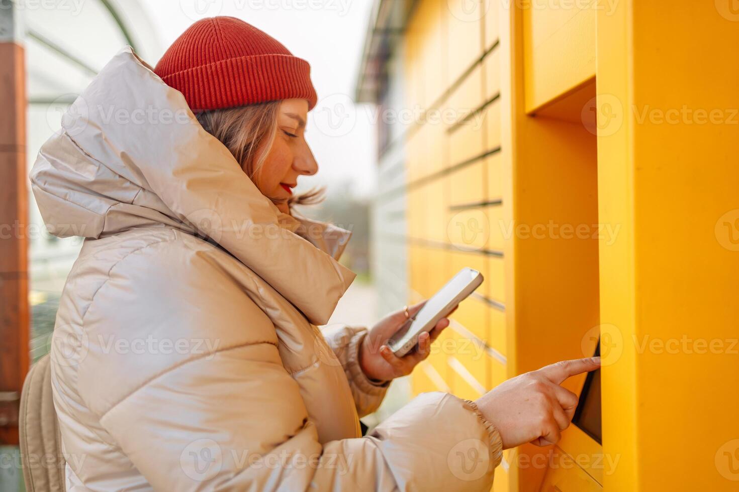 Smiling beautiful Caucasian woman client holding cardboard boxes and hands show smartphone using automated self service post terminal machine or locker. Mail shipping concept. photo