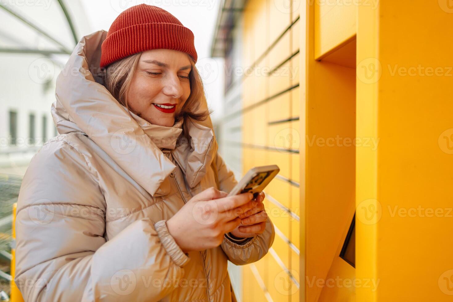 Happy smiling woman client with box at outdoor automated parcel machine choosing operation on touch screen by smart phone photo