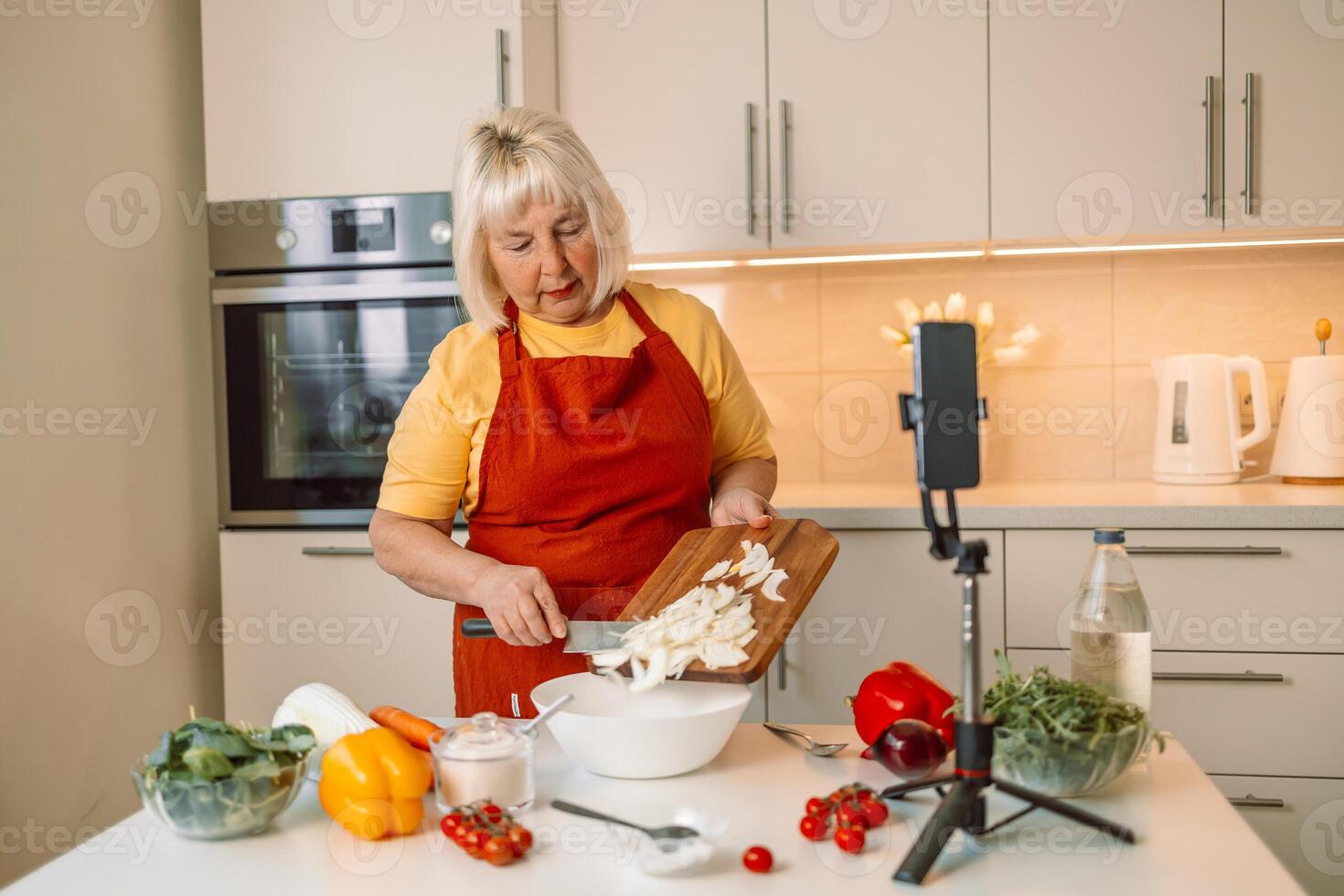 Hands of senior 60s female chopping fresh onion on wooden board while preparing italian pasta with vegetables. Caucasian female food blogger working on a new and explaining how to cook a dish photo