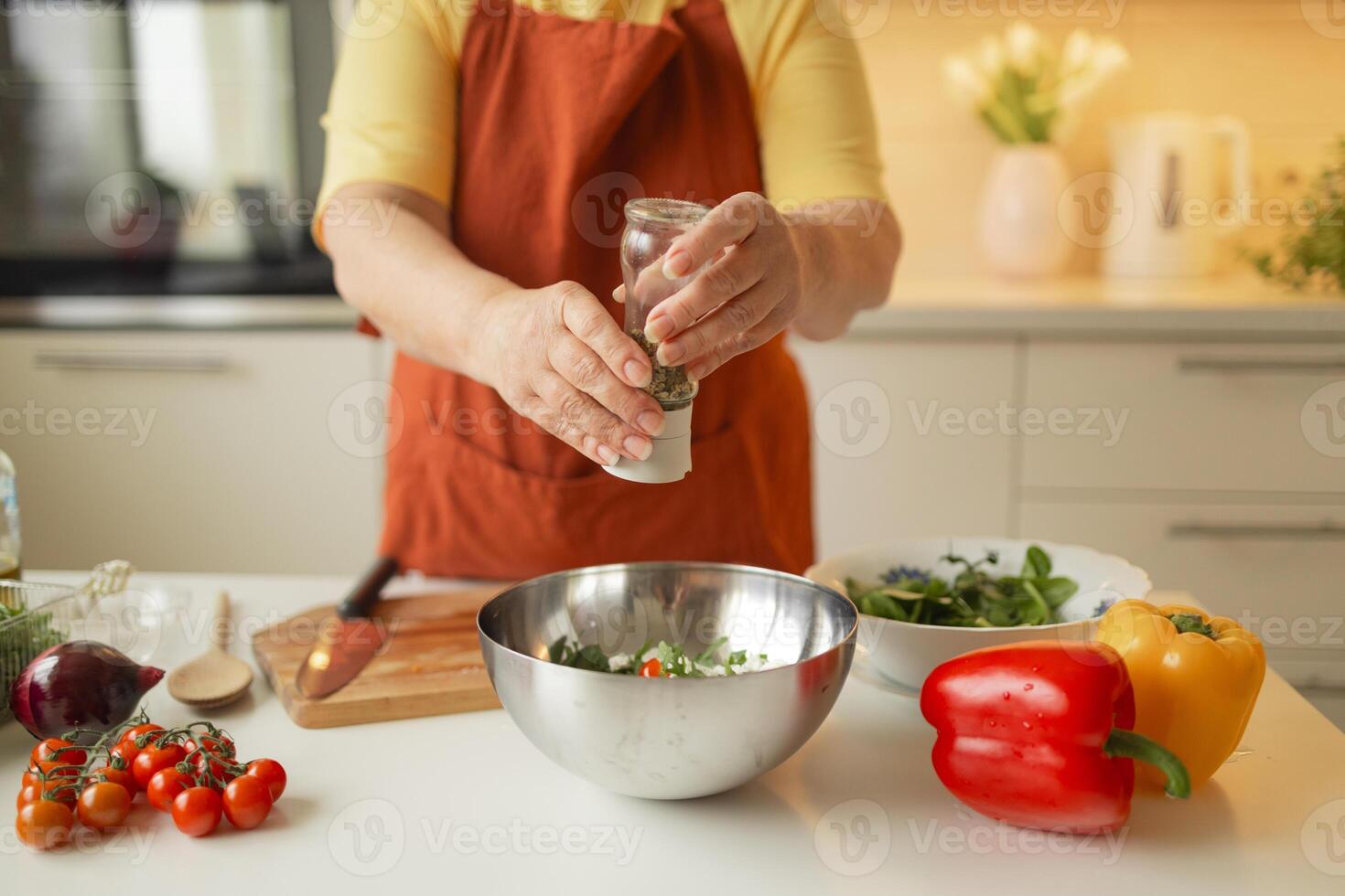 Close-up of a 60s woman preparing healthy vegetable salad in kitchen, seasoning meal, adding some herbs and salt to bowl, enjoying cooking vegetarian food at home, free space for advertising text photo