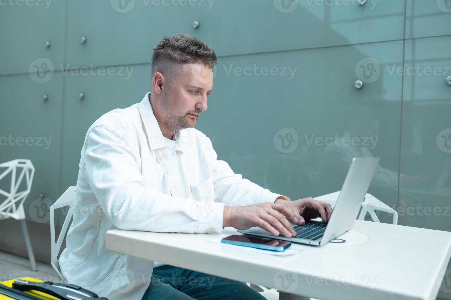 Happy traveling Caucasian man in stylish clothes uses mobile smartphone and laptop, checking trip destination on Internet. Male waiting at flight gates for plane boarding, smiling have a vacation trip photo