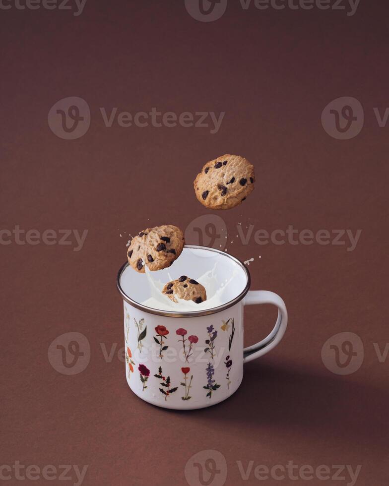 Chocolate chip cookies falling into vintage floral enamel mug full of fresh milk on brown background. Sweet food biscuit concept. Minimal layout. Trendy chocolate cookies with milk composition. photo