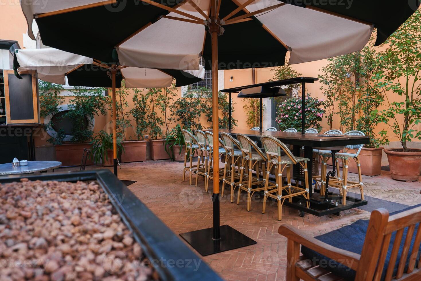 Luxury hotel outdoor seating area in Rome. Warm ambiance with terracotta tiles, cream umbrellas, rattan chairs, lush plants. Elegant and tranquil setting. photo